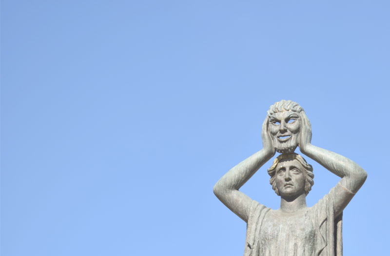 Image shows a statue of a person who is holding up a mask and revealing a true face